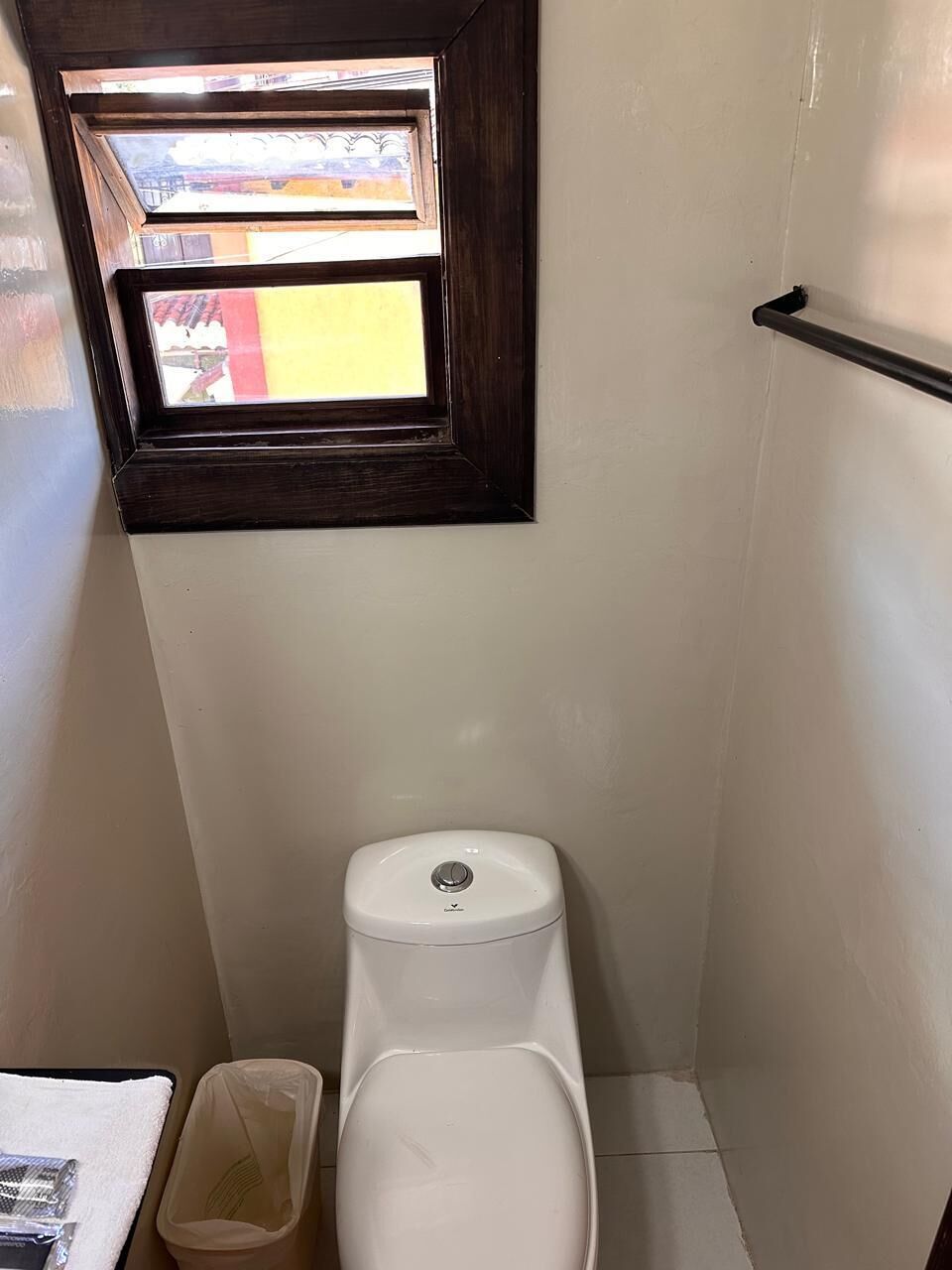 A small, narrow bathroom with a toilet, a window, and a trash bin.