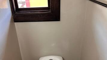 A small, narrow bathroom with a toilet, a window, and a trash bin.
