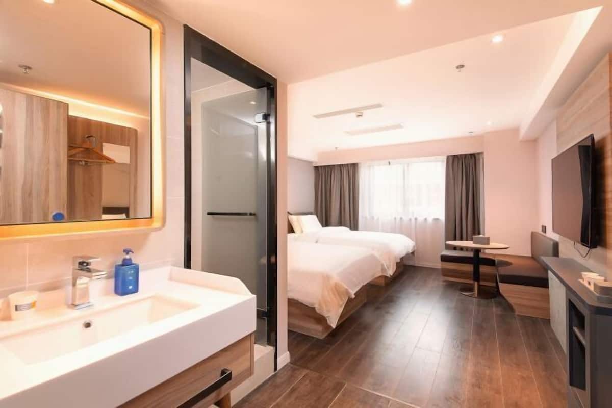 Una habitación de hotel con un espejo grande, un lavabo blanco, una cama, un televisor y una mesita pequeña.