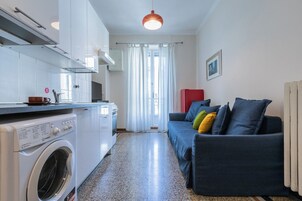 Un ambiente compatto con angolo cottura, lavatrice, divano con cuscini colorati e termosifone.