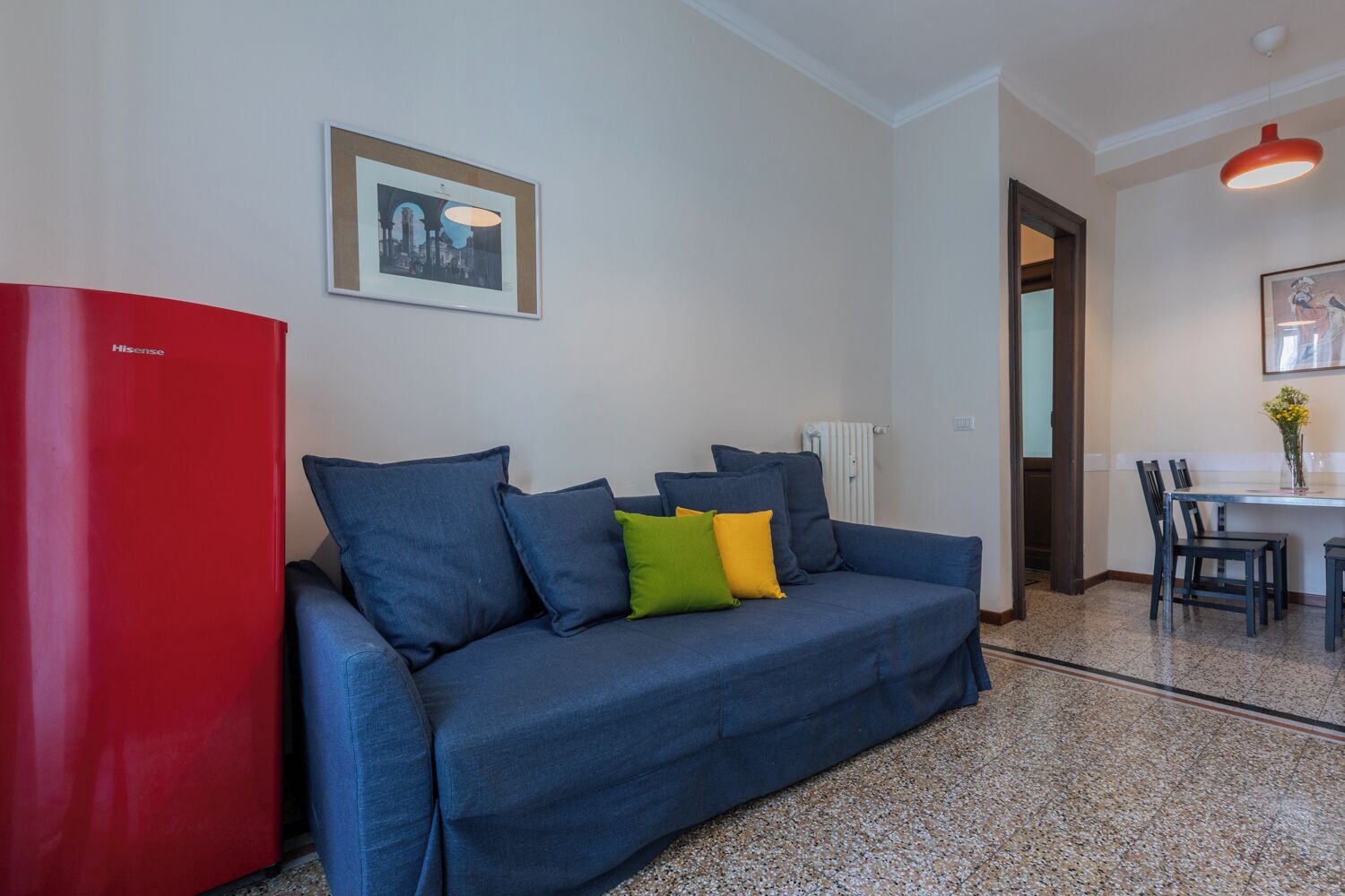 Ein blaues Sofa mit bunten Kissen, eine rote Kommode und ein gerahmtes Bild an der Wand.