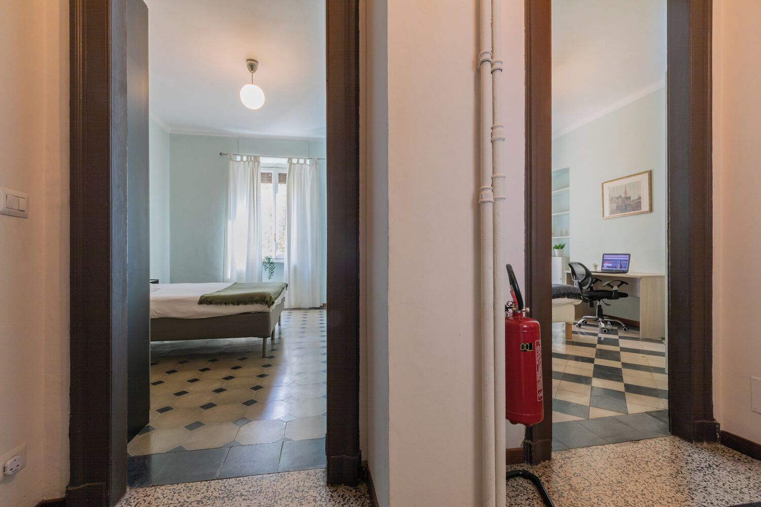 Una habitación de hotel con una cama, un escritorio con una computadora, un extintor y un piso a cuadros.