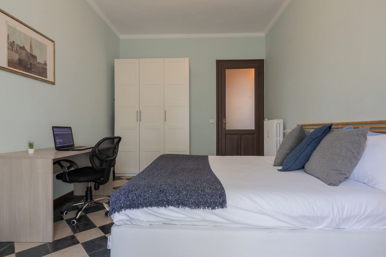 A bedroom with a bed, a desk with a laptop, a chair, a wardrobe, and a radiator.