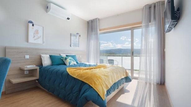 A modern hotel room with a large bed, a bedside table, a mounted TV, and a window with a view of the landscape. - Refúgio da Garça (Chaves)