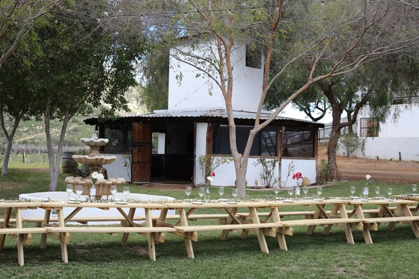 A rustic outdoor dining area with wooden picnic tables set up for a meal, surrounded by trees and a building with a white exterior. - Casa Luna Viñedo (Valle de Guadalupe)