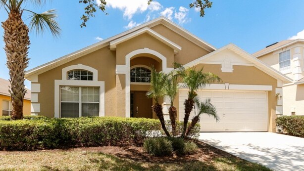 A two-story house with a beige facade, a garage, and a well-maintained front yard with palm trees and shrubs. - Beautiful 4 Bedroom Villa near Disney World with Private Pool (Kissimmee)