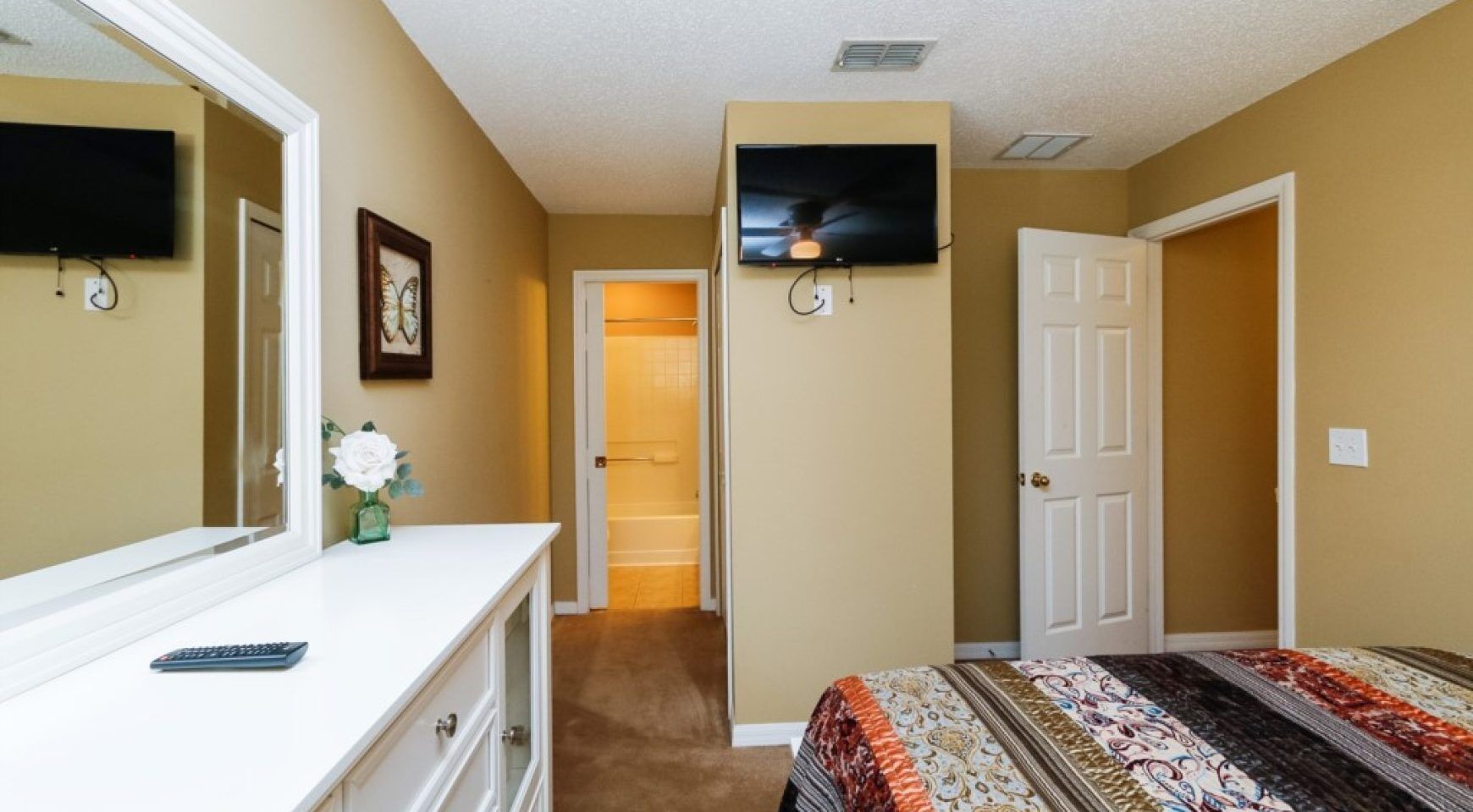 Une chambre avec un grand lit, une commode, un miroir, un téléviseur fixé au mur et une porte donnant sur un couloir.
