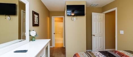 Une chambre avec un grand lit, une commode, un miroir, un téléviseur fixé au mur et une porte donnant sur un couloir.