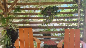 Eine Holzpergola mit zwei orangefarbenen Stühlen und einem kleinen Tisch, umgeben von üppiger Bepflanzung und einem gemusterten Teppich.