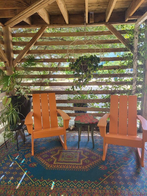 Een houten pergola met twee oranje stoelen en een kleine tafel, omringd door weelderige begroeiing en een vloerkleed met patroon.