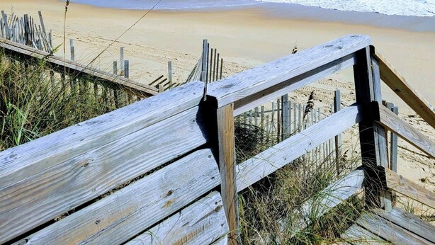 Ein hölzerner Steg führt zu einem Strand, wo Wellen an die Küste branden, unter einem wolkenlosen, blauen Himmel.