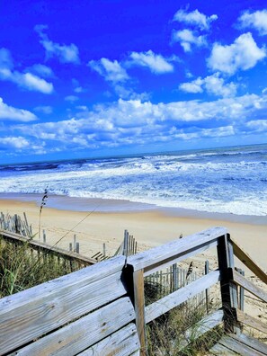 A wooden boardwalk leading to a beach with waves crashing onto the shore under a clear blue sky. - Steps from the beach in the heart of Duck! (Kitty Hawk)