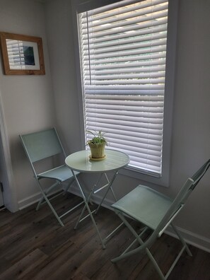 Ein Zimmer mit einem kleinen runden Tisch, zwei grünen Stühlen, einer Topfpflanze und einem Fenster mit weißen Jalousien.