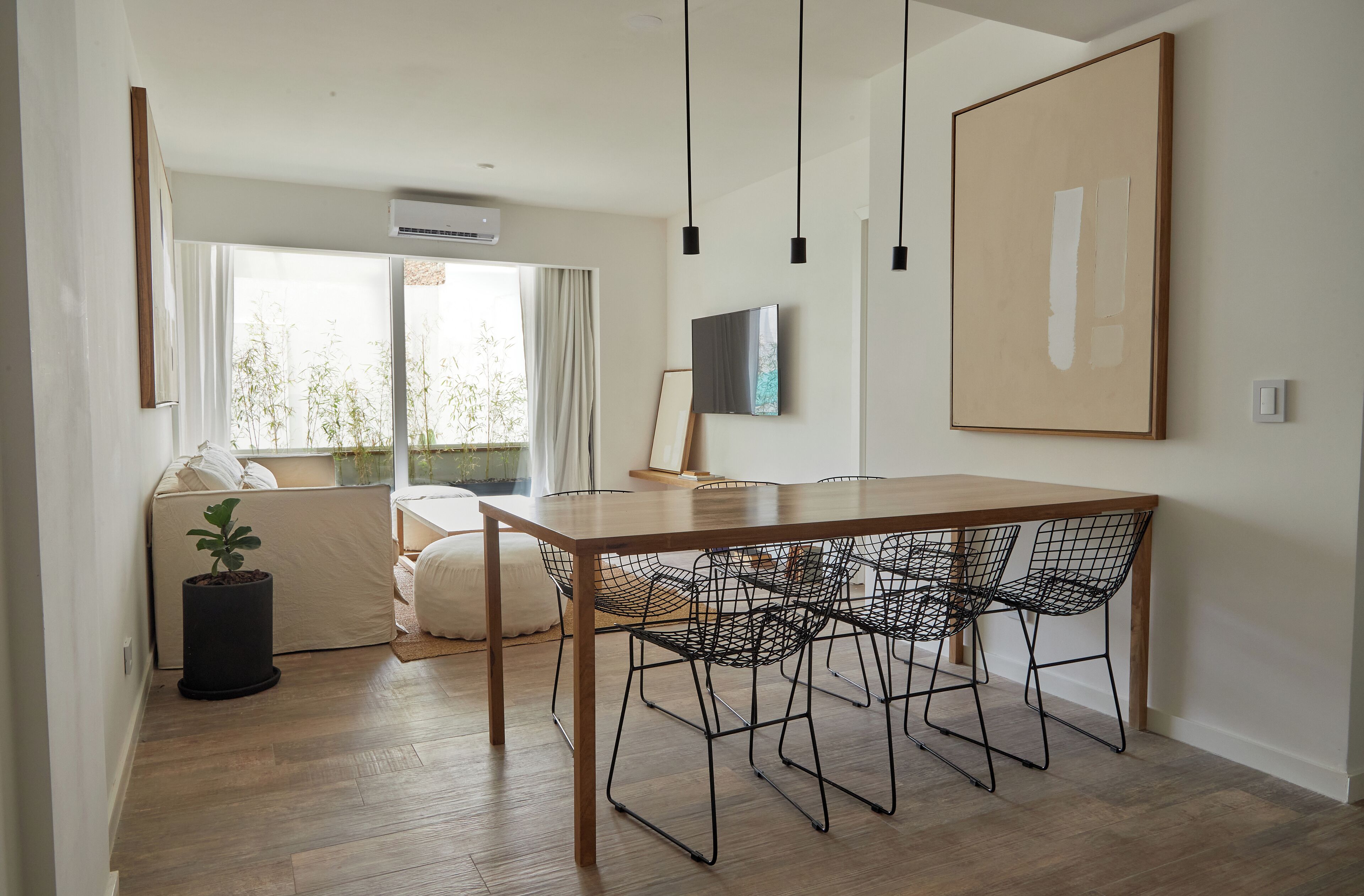 A modern dining area with a wooden table and wireframe chairs, a large framed artwork on the wall, and a window with curtains.