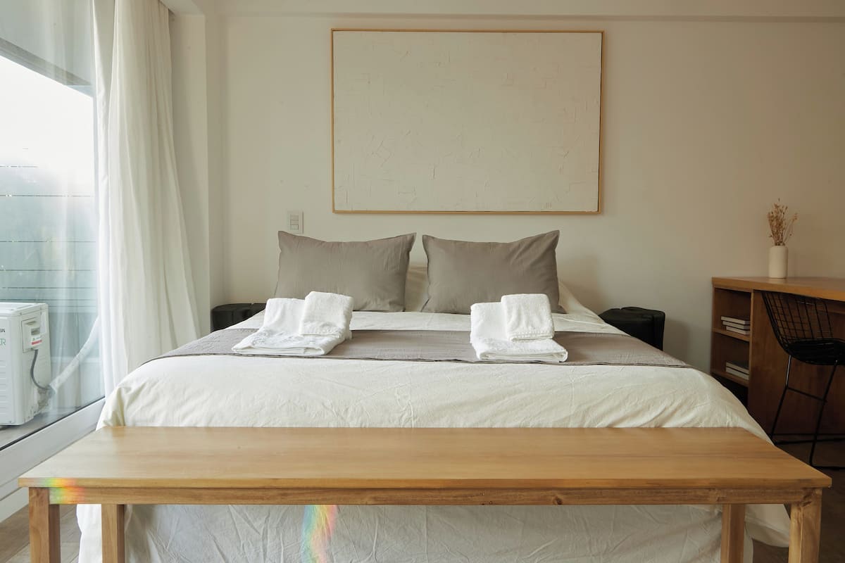 A hotel room with a bed, two pillows, a bedside table, a chair, a desk, a window with curtains, and a wall-mounted artwork.