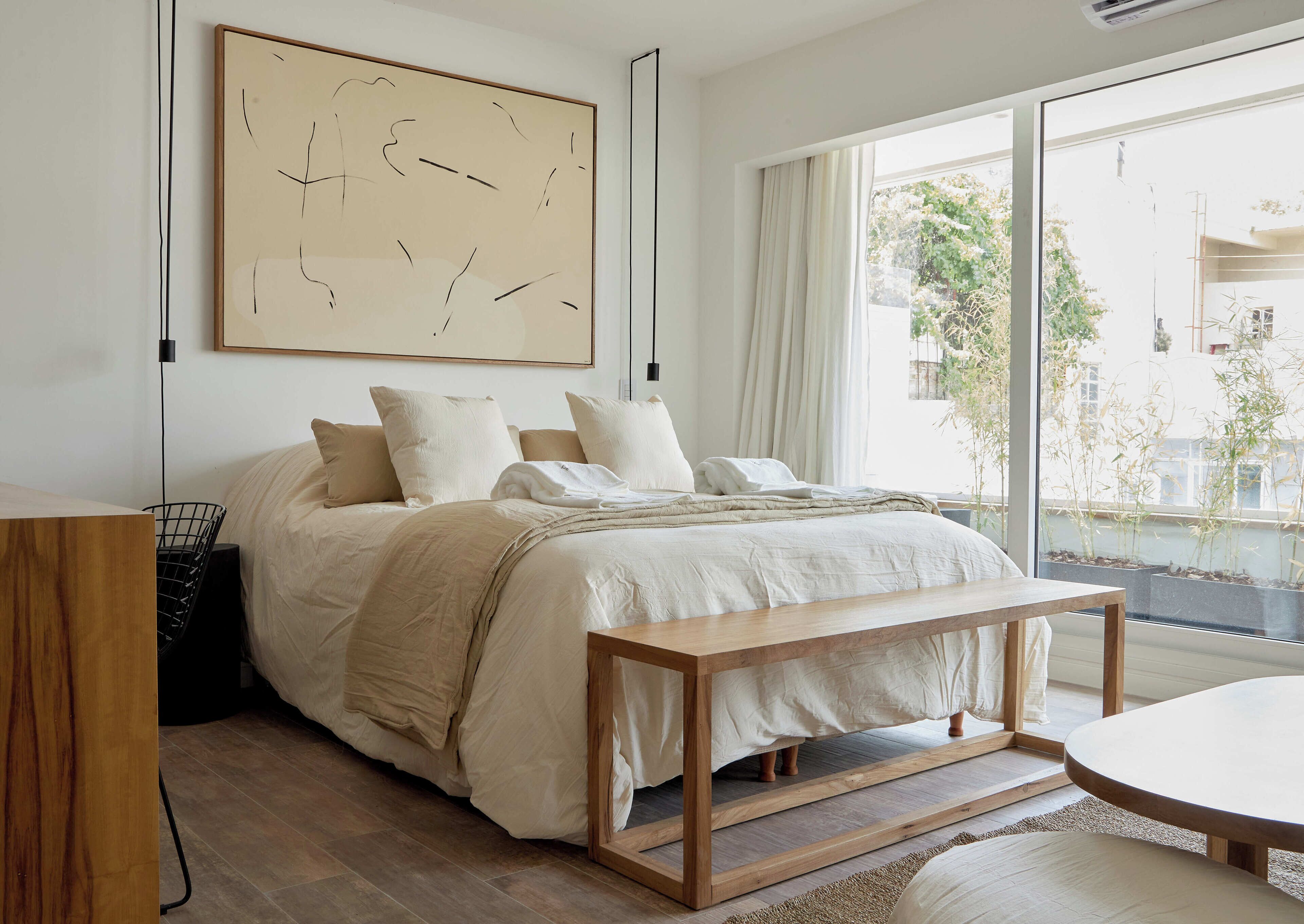 A modern bedroom with a large bed, a wooden bench, a large window with a view of the city, and a minimalist abstract painting on the wall.