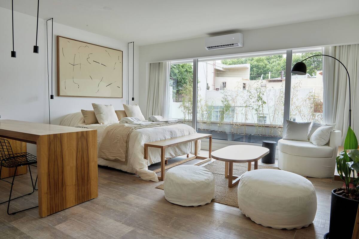 A modern bedroom with a bed, bedside table, bench, ottoman, and a view of the outside.