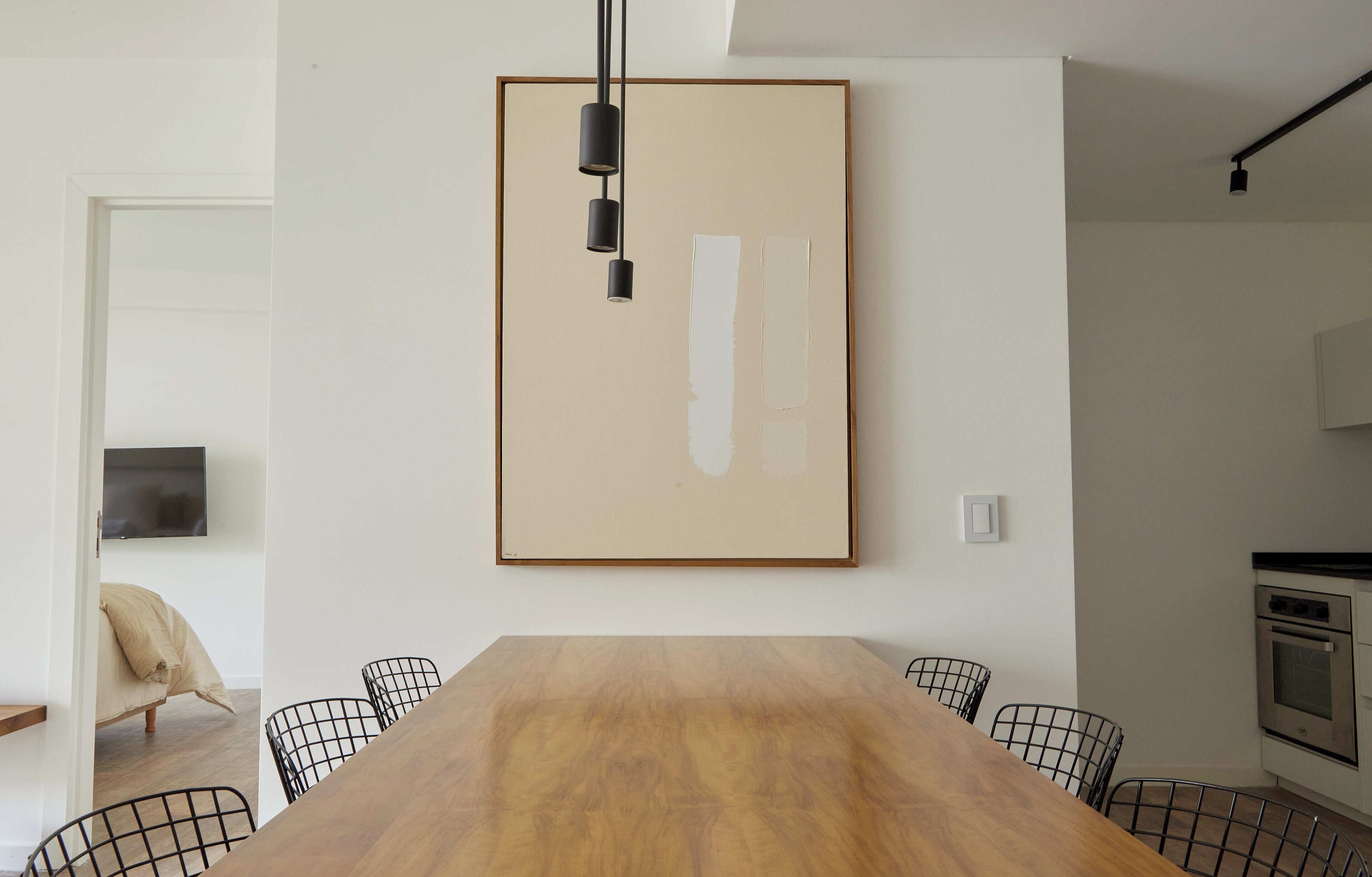 A dining area with a wooden table, black chairs, a framed abstract painting, and a kitchen in the background.