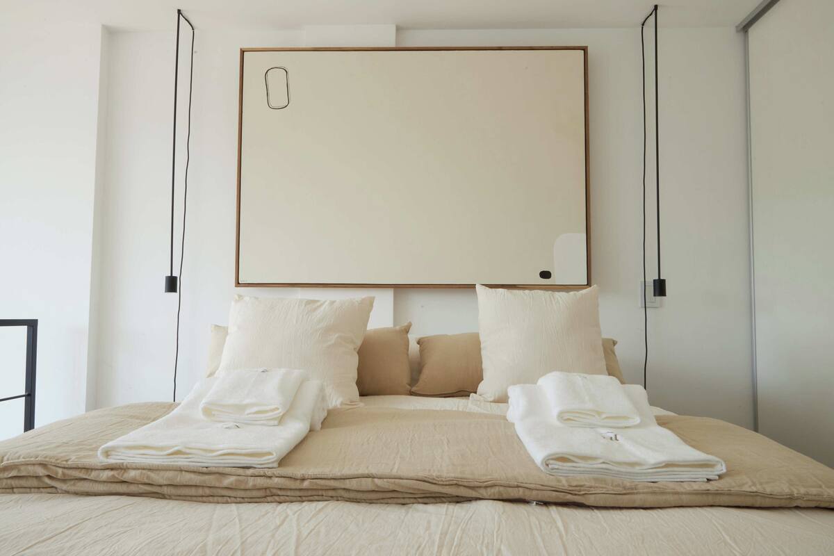 A neatly made bed with beige bedding and pillows, a large blank framed artwork above it, and a simple, clean room design.