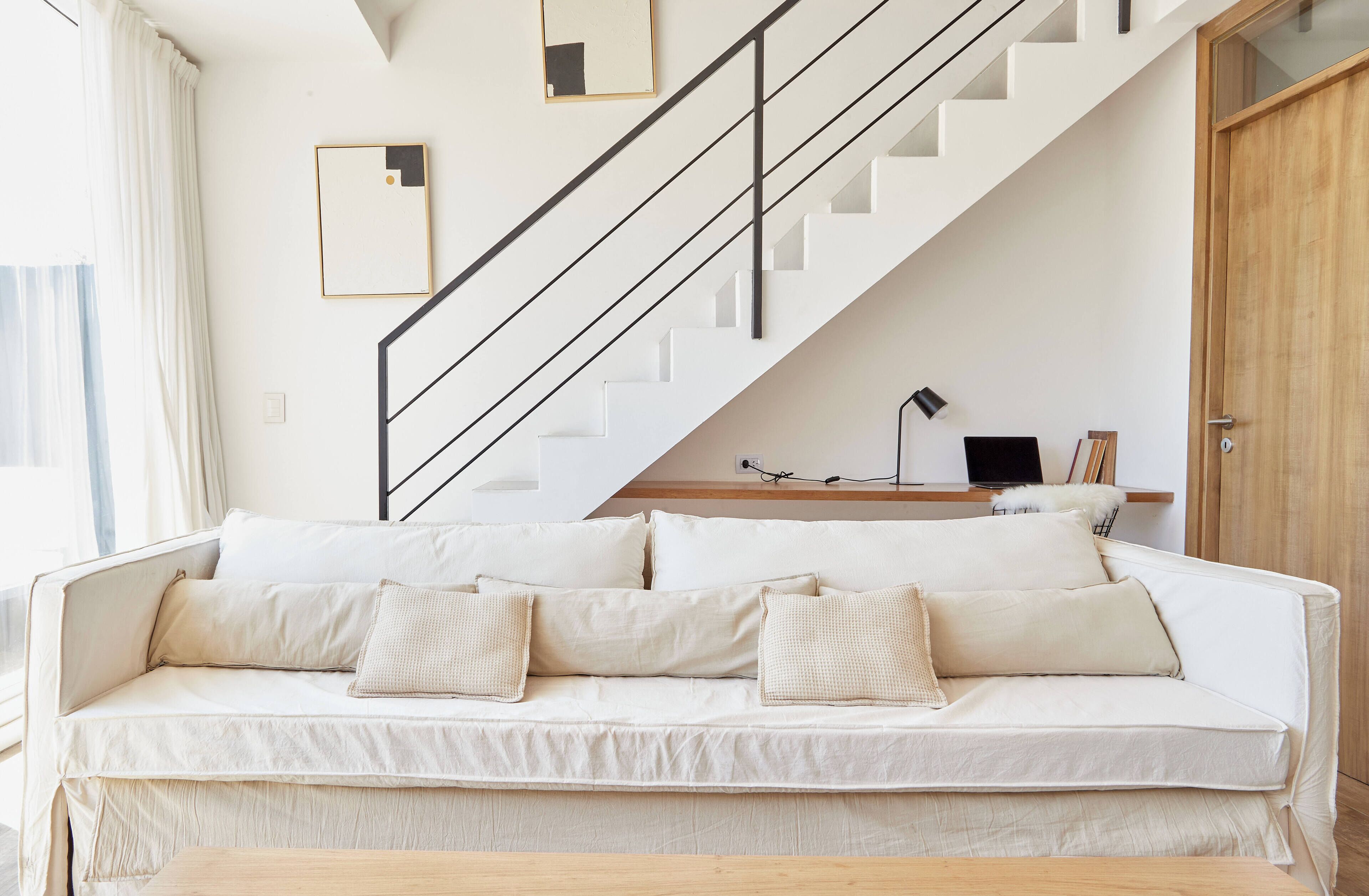 Una sala de estar moderna con un sofá blanco, una mesa de centro de madera, una escalera con barandilla negra y una puerta de madera.