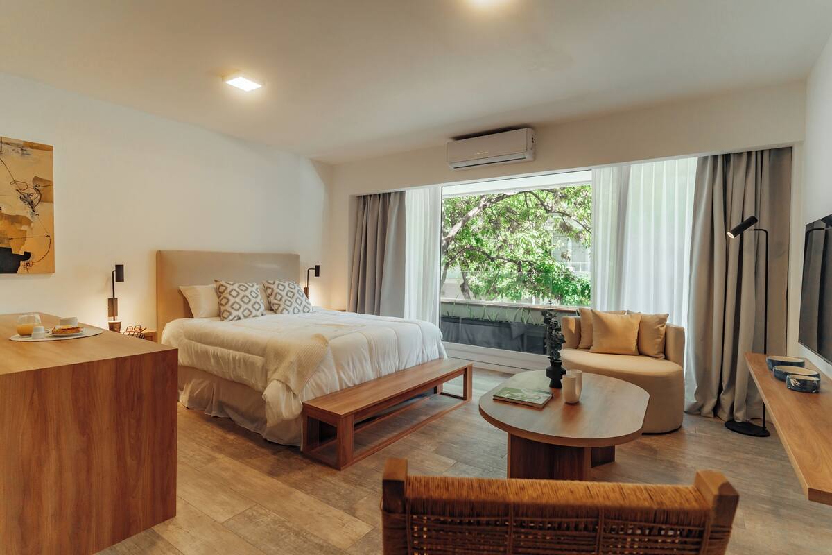 A modern hotel room with a large bed, a sofa, a dining table, and a TV.