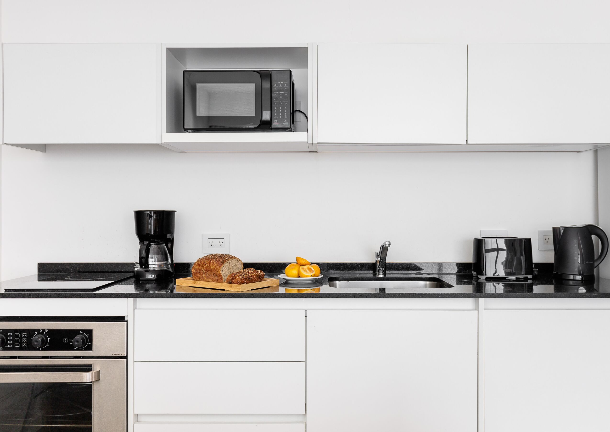 A modern kitchen with white cabinets, a built-in microwave, a coffee maker, a toaster, and a bread loaf on the counter.