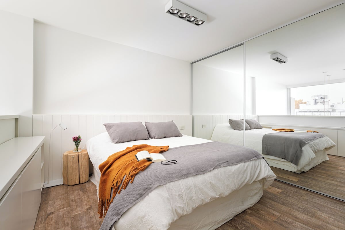 A modern bedroom with a large mirror, a bed with a gray and white duvet, a small wooden side table with a vase of flowers, and a window with a city view.