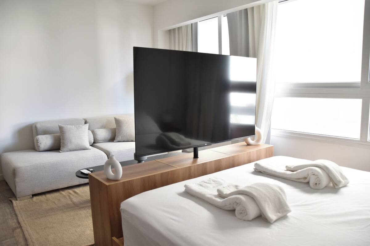 A modern hotel room with a large flat-screen TV, a sofa, a wooden bedside table, and neatly folded towels.