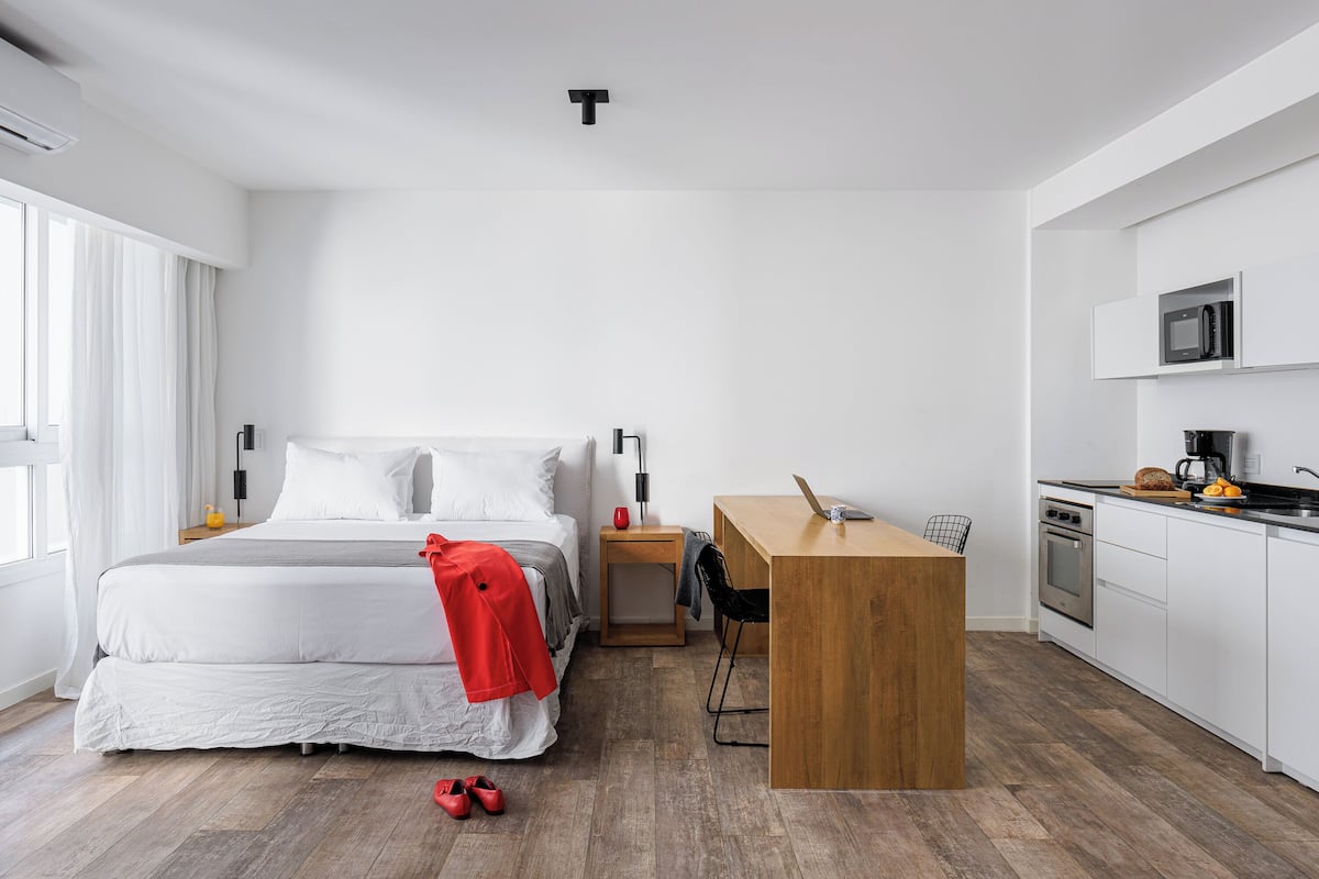 A modern hotel room with a large bed, a wooden desk, a kitchenette, and a view of the city.