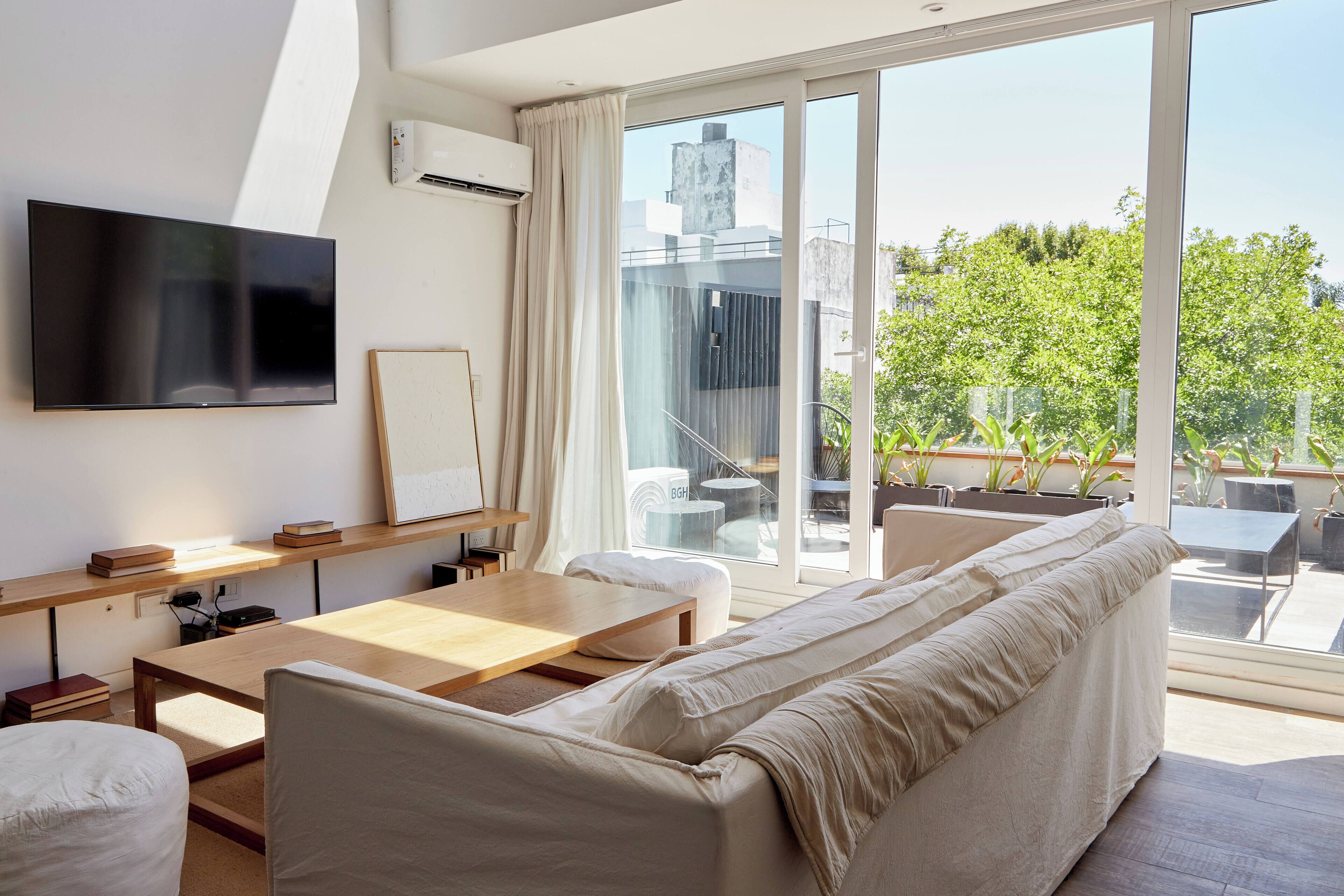A modern living room with a flat-screen TV, a wooden table, a sofa, and a balcony with a view of greenery and buildings.