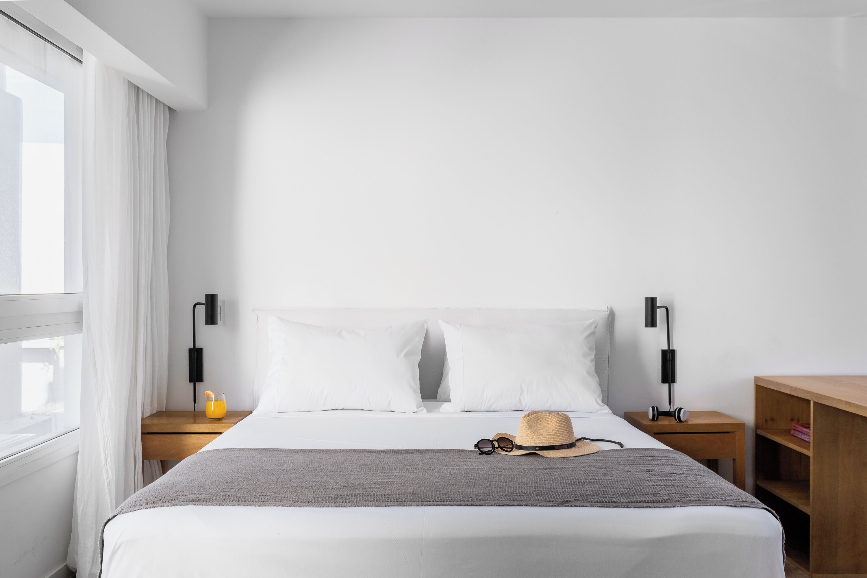 A neatly made bed with white bedding, a gray throw blanket, and a straw hat on the pillows. There are two bedside tables with black lamps and a small wooden nightstand with a shelf.
