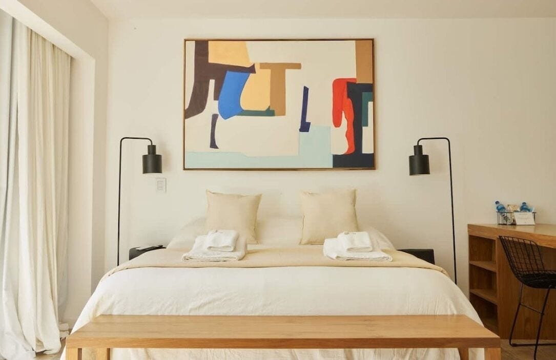 Habitación de hotel con cama, dos mesitas de noche, escritorio y una silla. En la pared, encima de la cama, hay un cuadro.