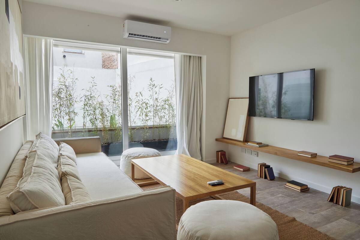 A modern living room with a sofa, a wooden coffee table, and a TV mounted on the wall. There are bookshelves with books and a potted plant near the window.
