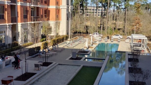Uma área de piscina com espreguiçadeiras e uma piscina pequena, cercada por um jardim bem cuidado e um edifício com vários andares.