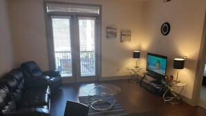 Uma sala de estar com um sofá de couro preto, uma mesa de jantar posta com pratos e copos, uma TV sobre um suporte e uma mesa de centro de vidro.