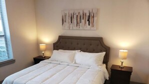 Cama bem arrumada com edredom branco, duas mesas de cabeceira com luminárias, e uma obra de arte na parede acima da cama.