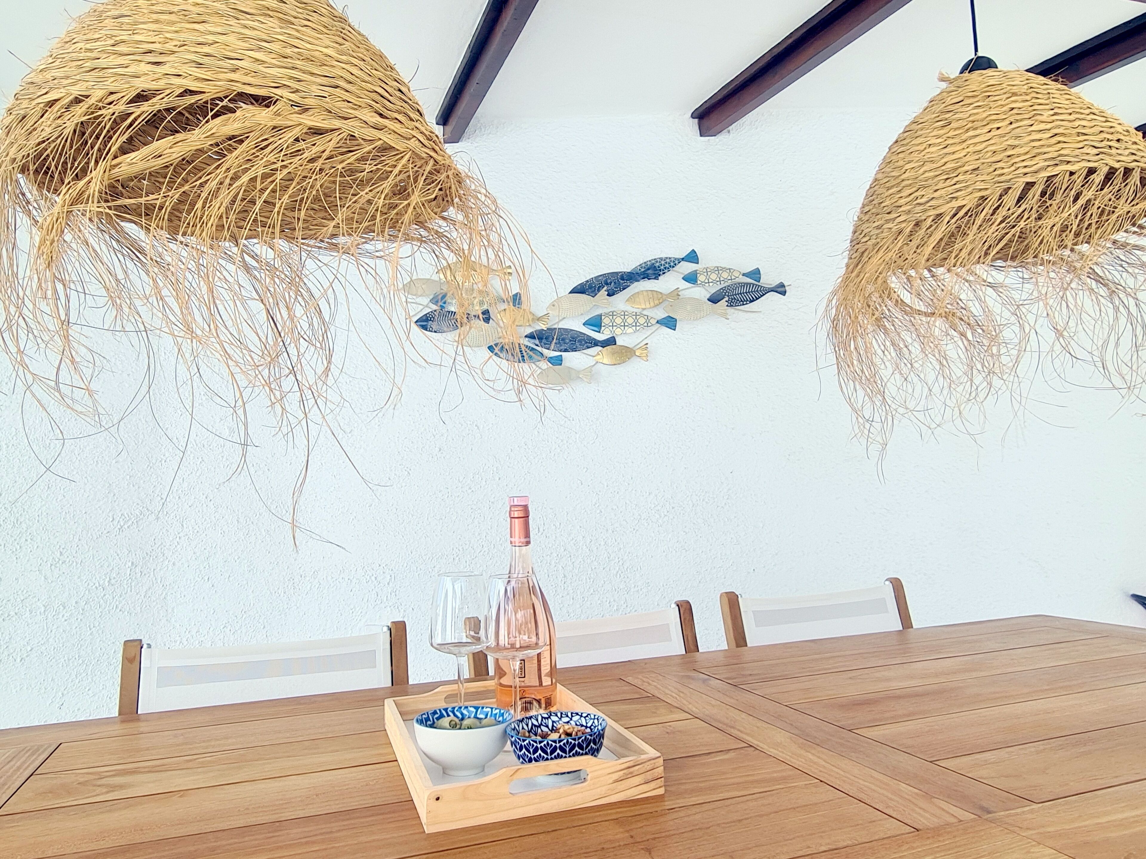 Un comedor con una mesa de madera, dos lámparas de mimbre colgantes, decoración de peces en la pared y una bandeja con una botella y copas.