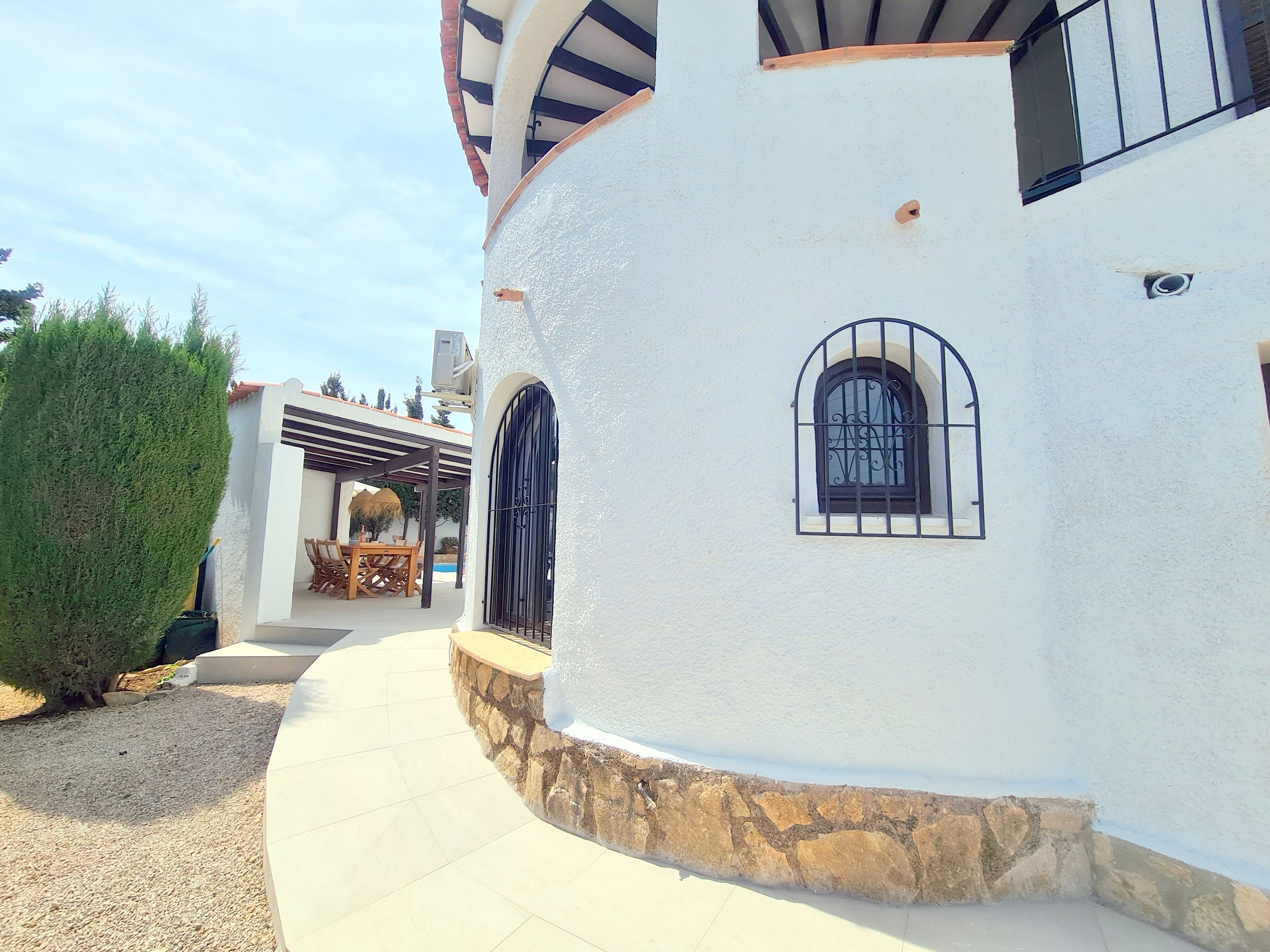 Un edificio blanco con base de piedra, un patio cubierto con una mesa y sillas, y una ventana pequeña con reja de metal.