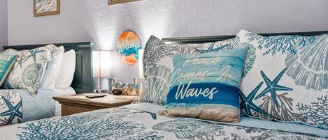 Un dormitorio con cama, cubrecolchón con motivos marinos en azul y blanco, almohadas con diseños náuticos y cuadros en las paredes.