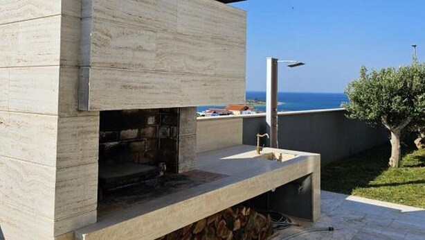 A modern outdoor kitchen with a stone fireplace, a built-in grill, and a view of the sea.