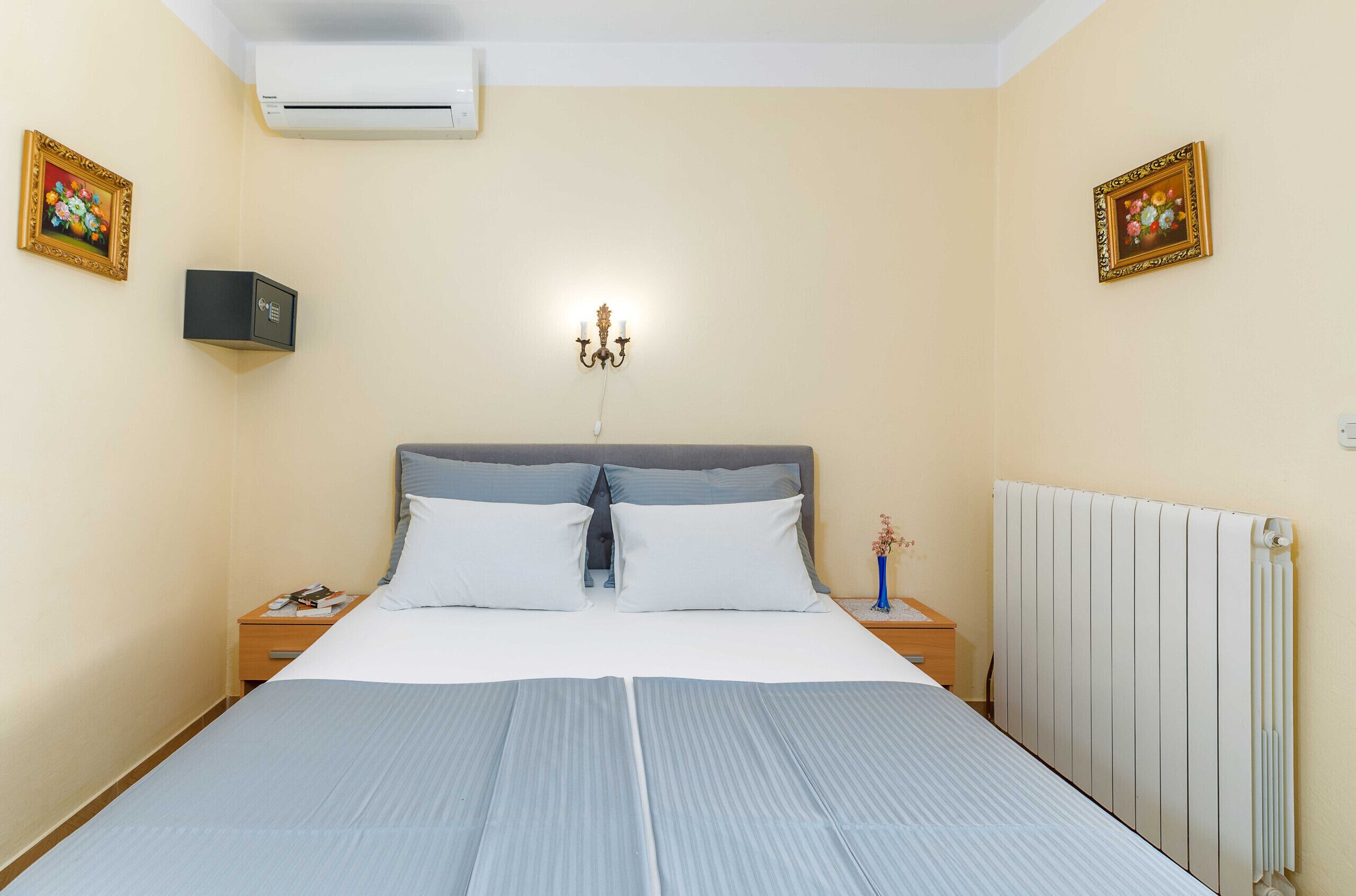 Una cama bien hecha con un cabecero gris, una mesita de noche con una lámpara y un aire acondicionado instalado en la pared.