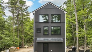 A two-story cabin with a dark exterior, surrounded by trees. - In the Trees - NH w/ Lake Access (Wakefield)