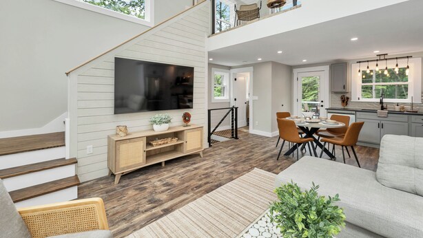 A spacious living area with a staircase leading to a balcony, a dining area with a kitchen in the background, and a large flat-screen TV mounted on the wall. - In the Trees - NH w/ Lake Access (Wakefield)