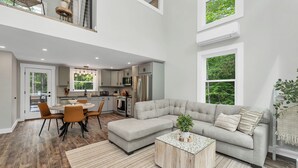 A spacious living area with a large grey sectional sofa, a wooden coffee table, and a dining area with a round table and orange chairs. - In the Trees - NH w/ Lake Access (Wakefield)