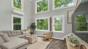 A spacious living room with a large sectional sofa, a wooden coffee table, and a dining table with chairs. There are multiple windows providing views of the outdoors. - In the Trees - NH w/ Lake Access (Wakefield)