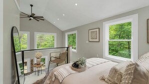 A bedroom with a large bed, a ceiling fan, and multiple windows offering a view of greenery. - In the Trees - NH w/ Lake Access (Wakefield)