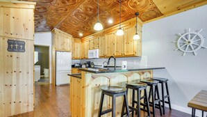 A kitchen with wooden cabinets, a central island with stools, a microwave, and a ship's wheel decoration. - Lakefront Vacation Rental ~ 10 Mi to Shipshewana! (Howe)