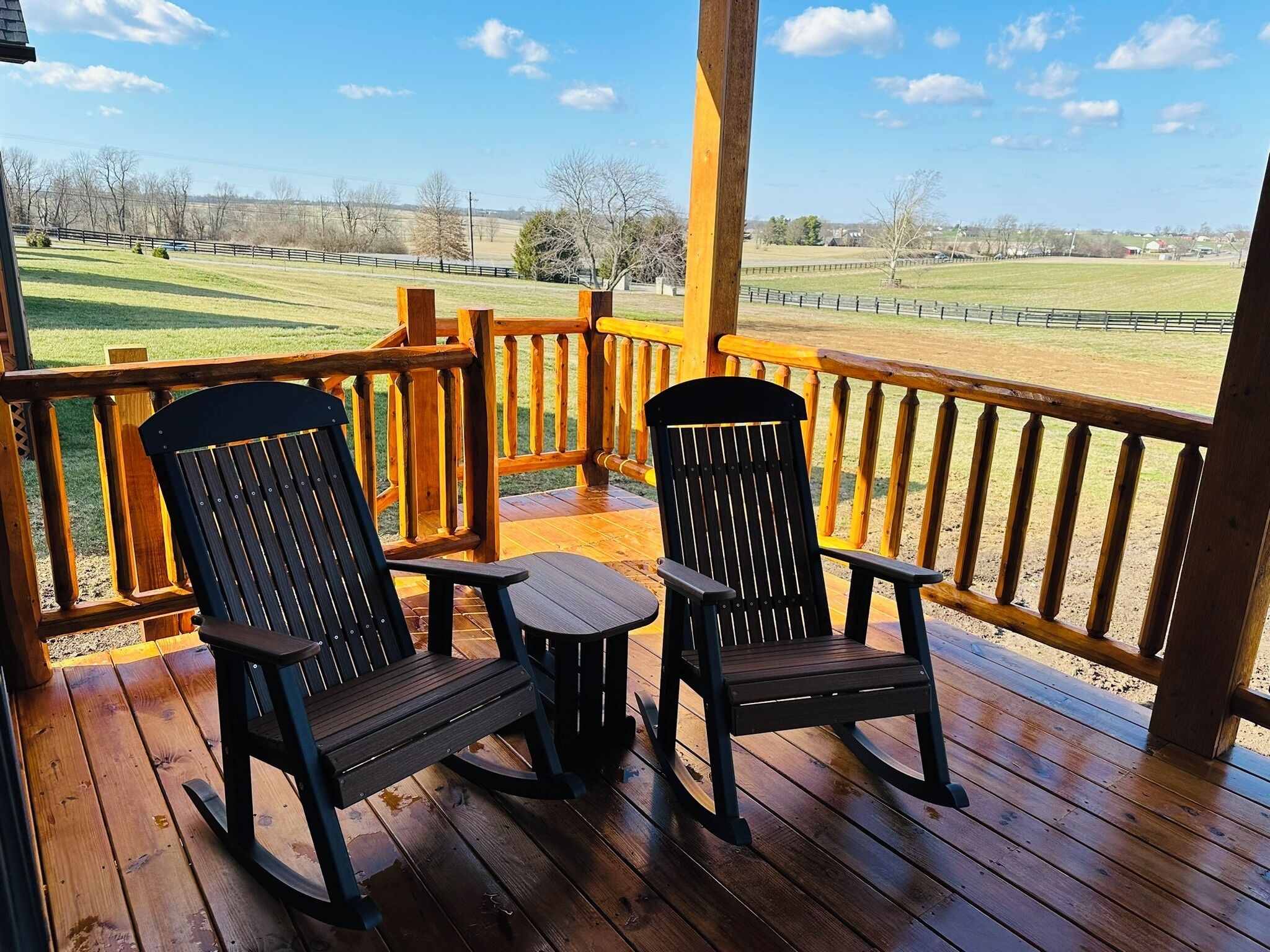 A wooden deck with two rocking chairs and a small table, overlooking a grassy field and distant buildings.