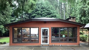 A small, single-story building with large windows and a door, surrounded by a paved area and dense greenery.