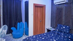 Una camera da letto con un letto, una poltrona blu, una ventola da terra, una porta in legno e un condizionatore d'aria.